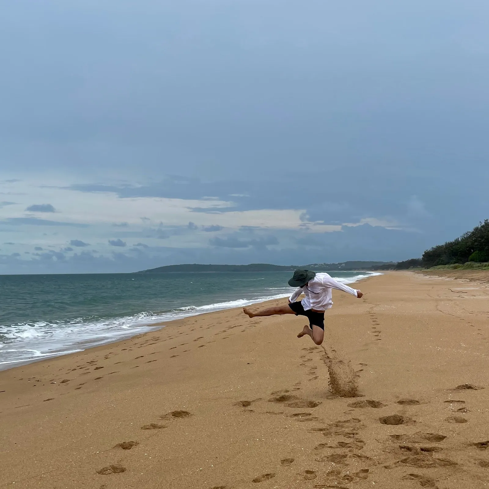 Paul mid-jump on an empty beach, kicking up sand, in a white shirt and a wide hat, with the sea to the left and a line of footprints behind him under a grey sky.